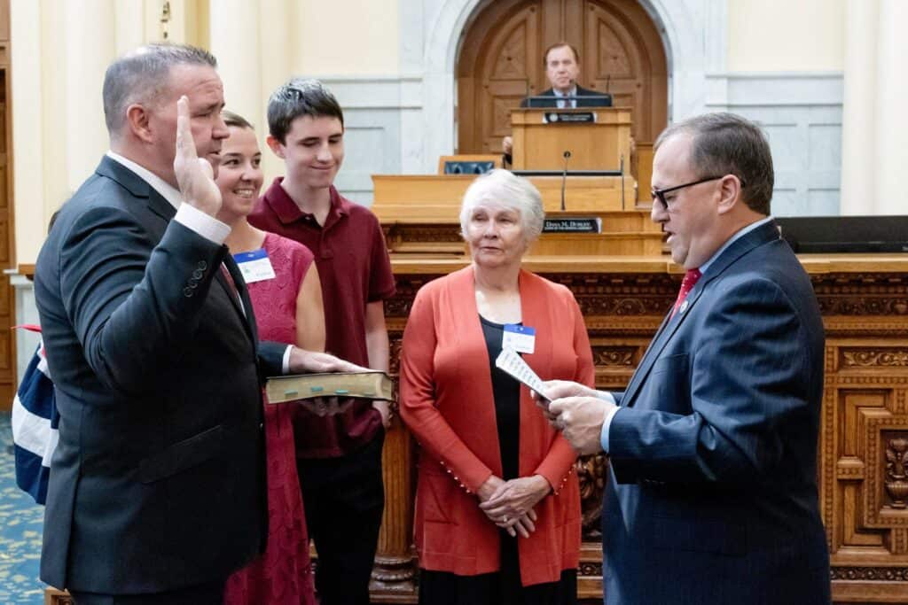 Alex Sauickie stands with his family, including wife, Maggie (holding 22-month-old daughter, Emma), and son, Alex, and the late Assemblyman Ron Dancer’s wife, Brenda, as Assembly Republican Leader John DiMaio swears him in from the floor of the Assembly chamber and Speaker Craig Coughlin watches from the dais.