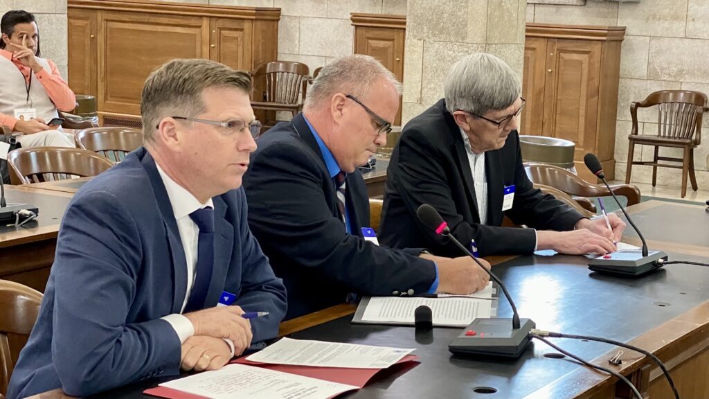 Three individuals sitting at a desk with microphones to give committee testimony.